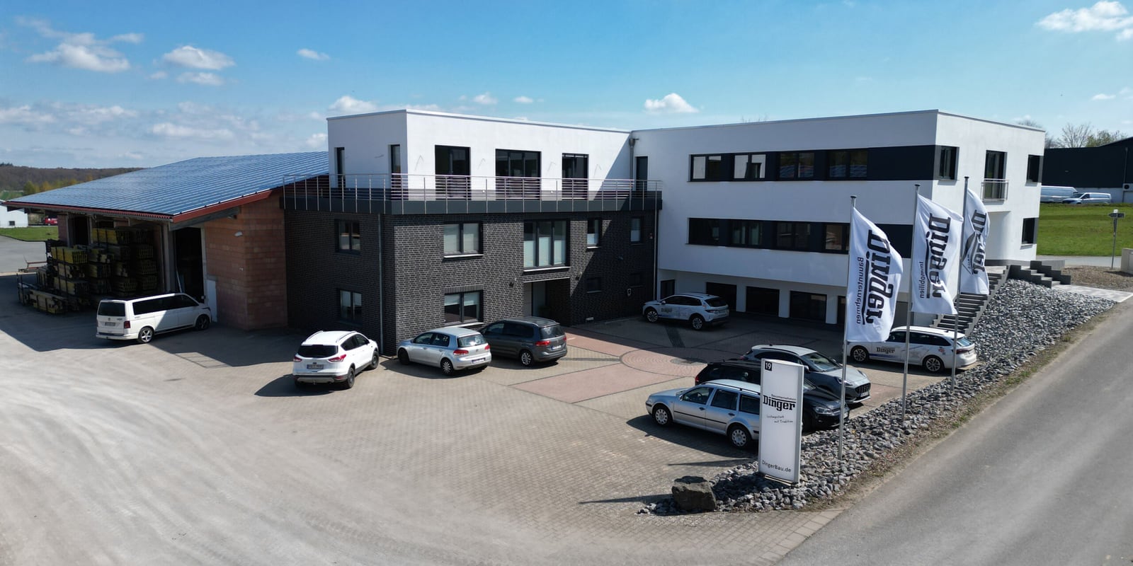 Luftaufnahme des Firmensitzes von Dinger Bauunternehmen mit Bürogebäude, Lagerhalle und Firmenfahnen