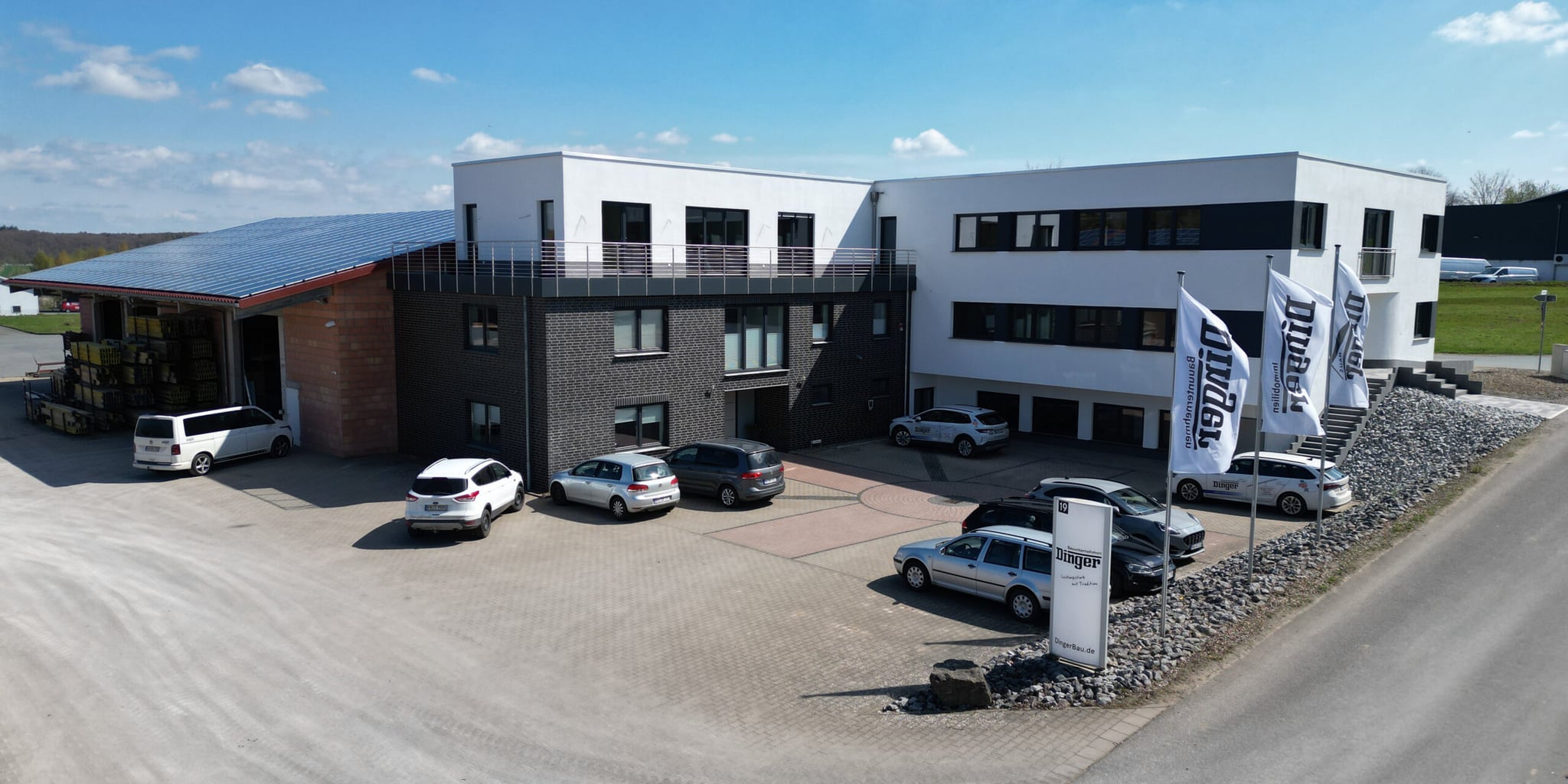 Luftaufnahme des Firmensitzes von Dinger Bauunternehmen mit Bürogebäude, Lagerhalle und Firmenfahnen
