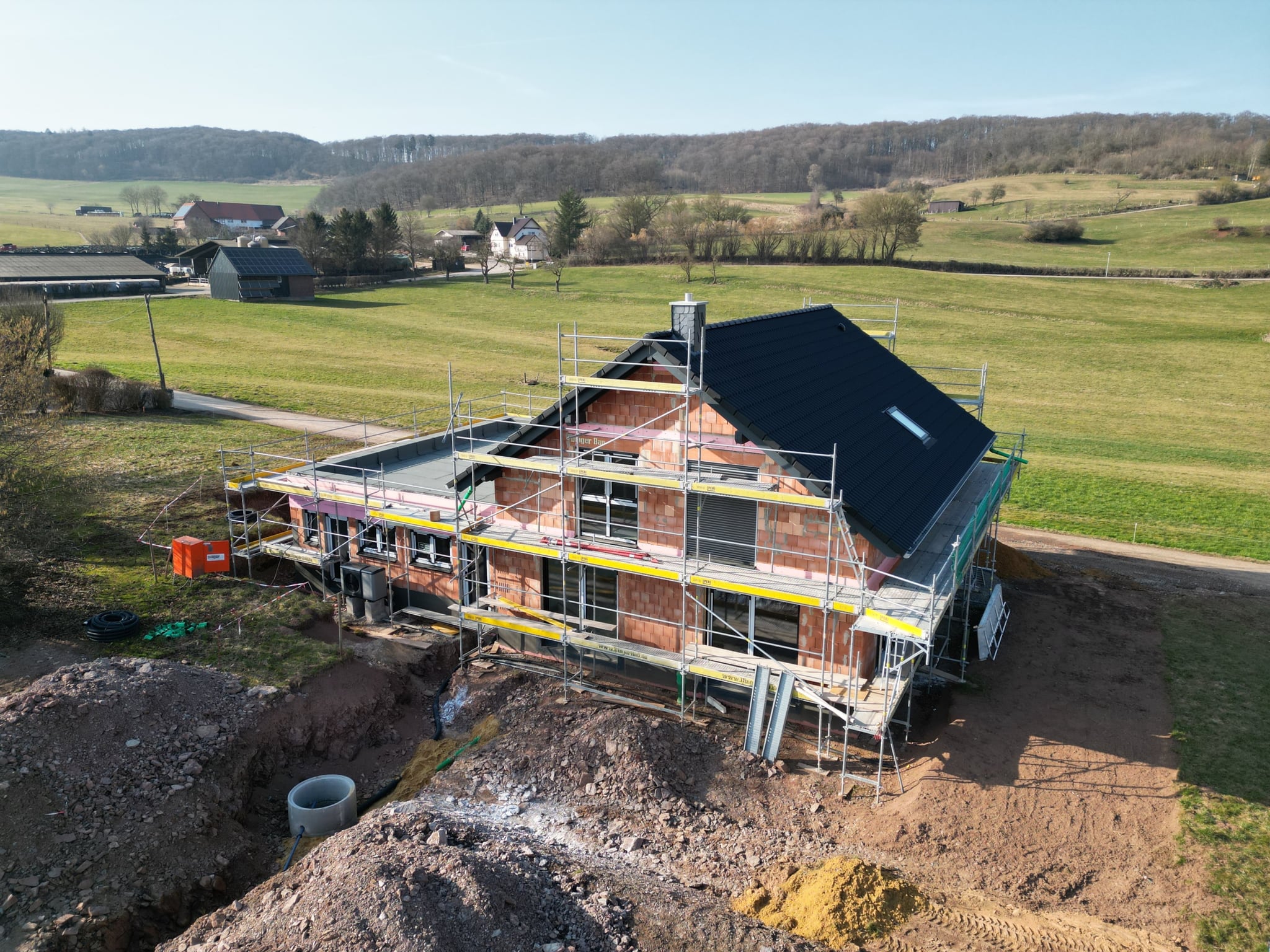 Eingerüsteter Rohbau mit fertig eingedecktem Dach aus der Luft, umgeben von Feldern und Wald