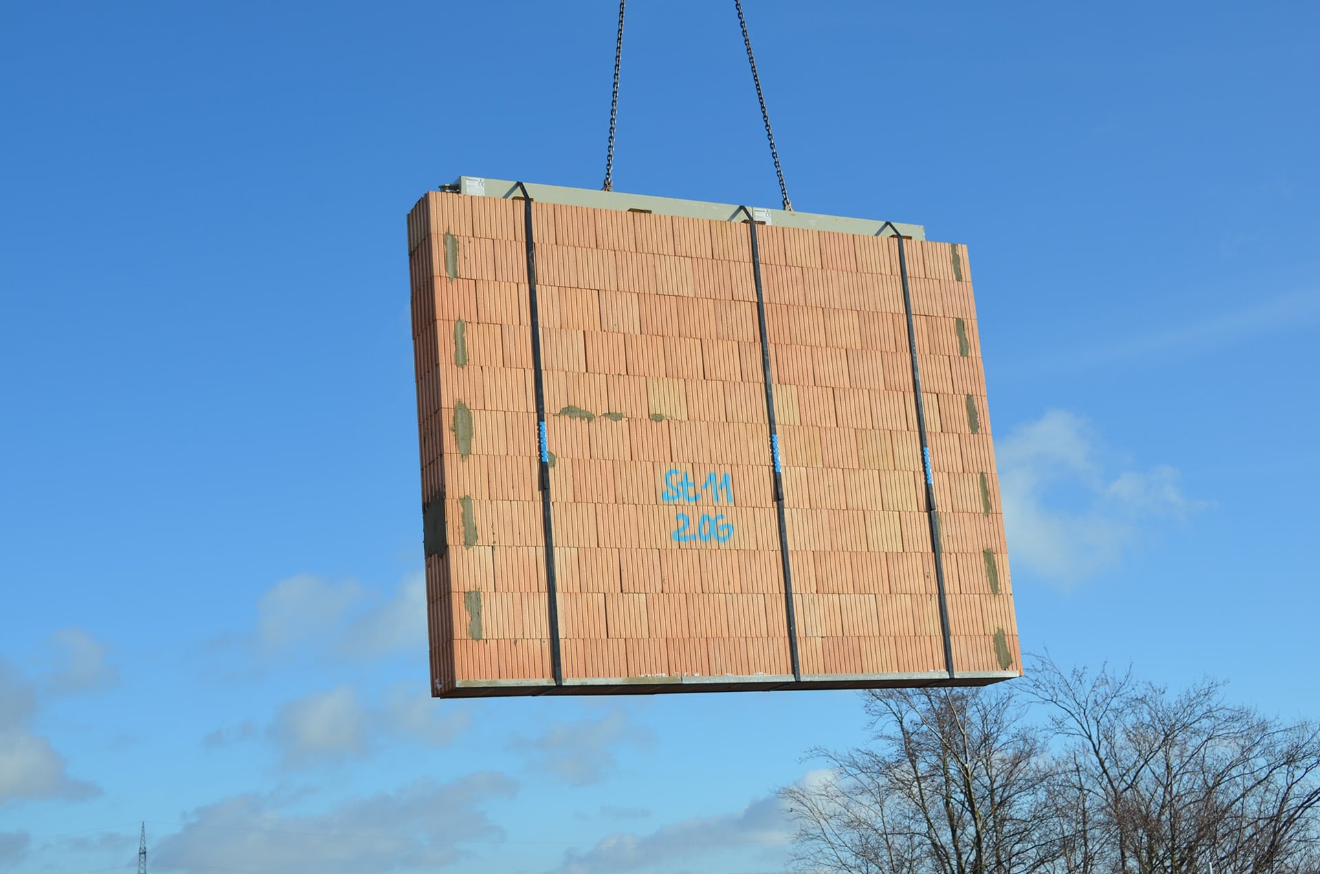 Eine Ziegel-Fertigteilwand hängt am Kranhaken über einer Baustelle